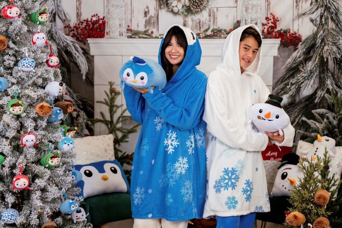 Two people wear snuggly soft fleece Peckington the Penguin blue and white hooded blankets with snowflake patterns in a festive, winter-themed room. One holds a penguin plush, the other a snowman plush. Plush toys decorate the tree and background.