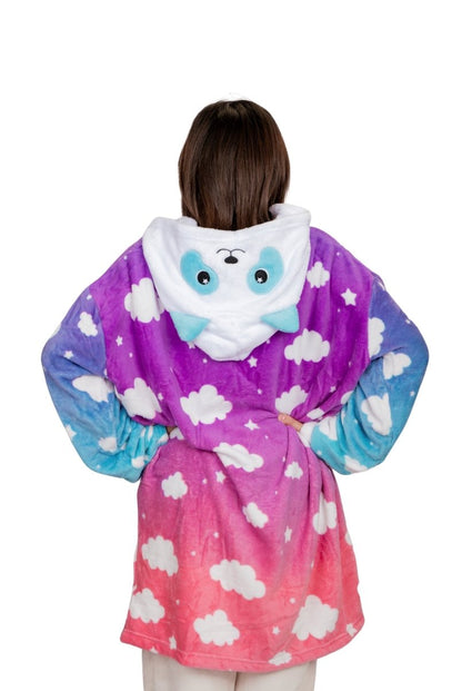 A person stands with their back to the camera, draped in a Panda Hooded Blanket featuring vibrant pink, purple, and blue cloud patterns. The hood resembles a plushie panda with an adorable face complete with big eyes and ears.