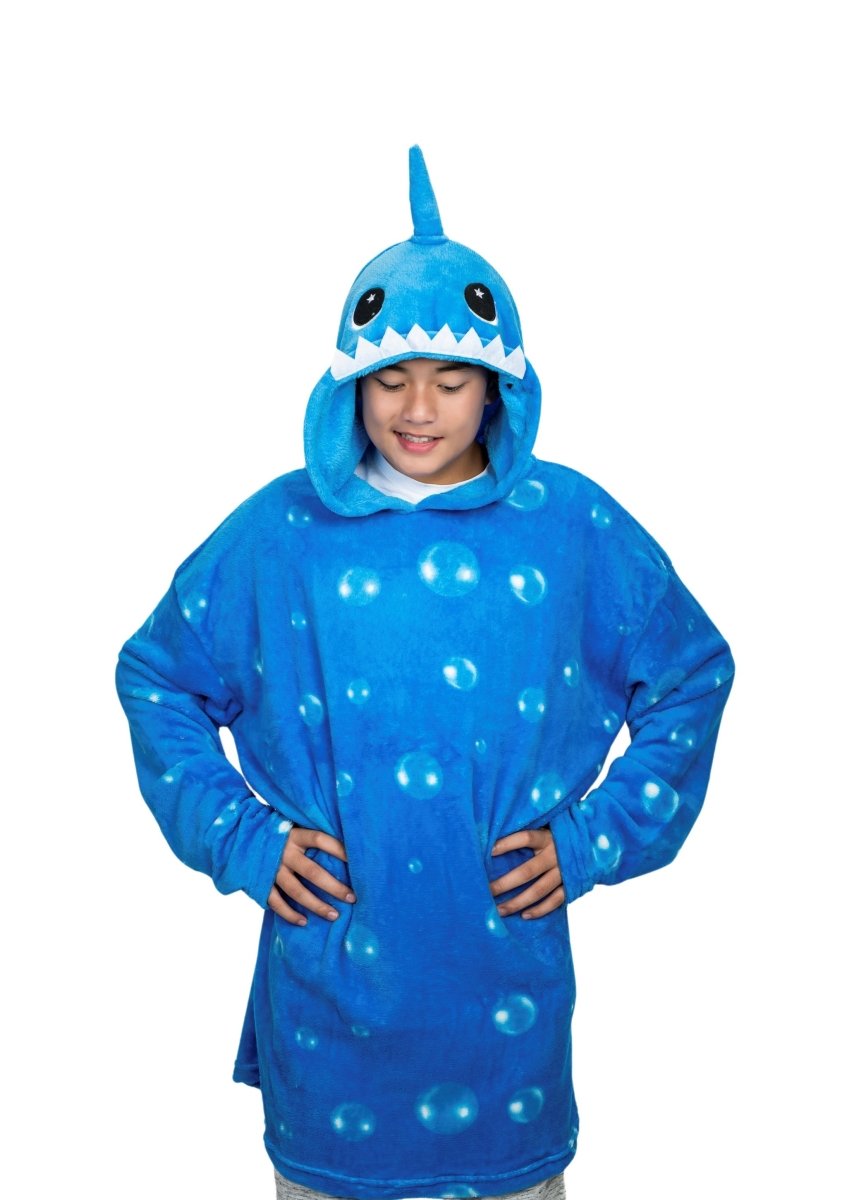 A person is wearing a Shark Hooded Blanket, an oversized plush blue hoodie with a shark fin on the hood and teeth around the face opening, decorated with soft bubble patterns like a beloved plush toy. Their hands rest on their hips.