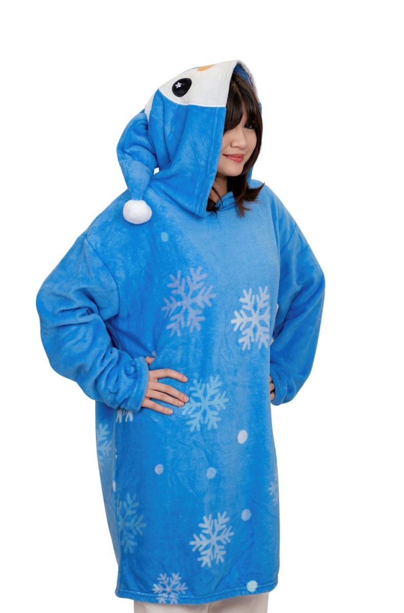A person stands with hands on hips and smiles, wearing the Peckington the Penguin Hooded Blanket—soft fleece in blue with white snowflakes and a penguin face on the hood.