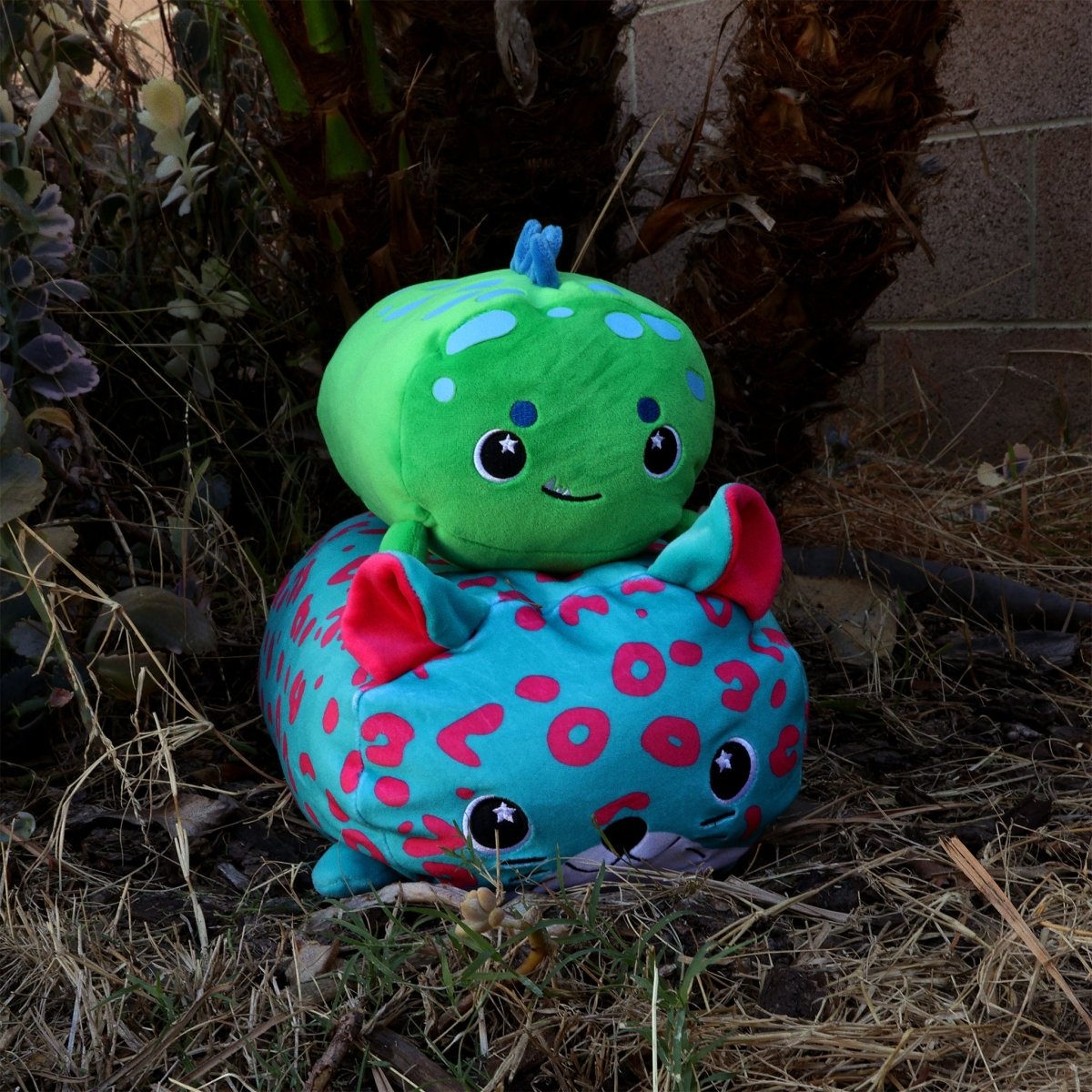 Two Sinclair the Dino plushies sit outside on dry grass. The green one with a blue stem and smile rests atop the blue one, which has pink spots, red ears, and a cute face. Plants can be seen in the background.