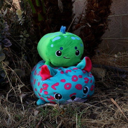 Two Sinclair the Dino plushies sit outside on dry grass. The green one with a blue stem and smile rests atop the blue one, which has pink spots, red ears, and a cute face. Plants can be seen in the background.