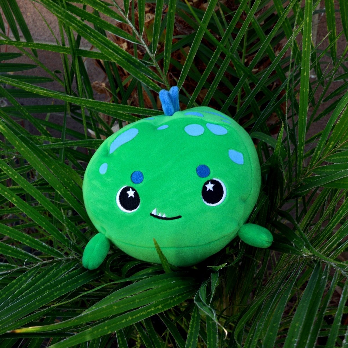 Sinclair the Dino, a round green plushie with a cute smiling face, blue spots, and small arms, sits nestled among green palm leaves.