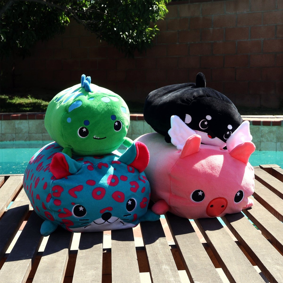 Four cuddly plush toys, including Barnaby the Flying Pig with wings, a soft stuffed cat, and two other vibrant creatures, are stacked in two rows on a wooden table by a pool. Sunlight and a brick wall provide warmth in the background.