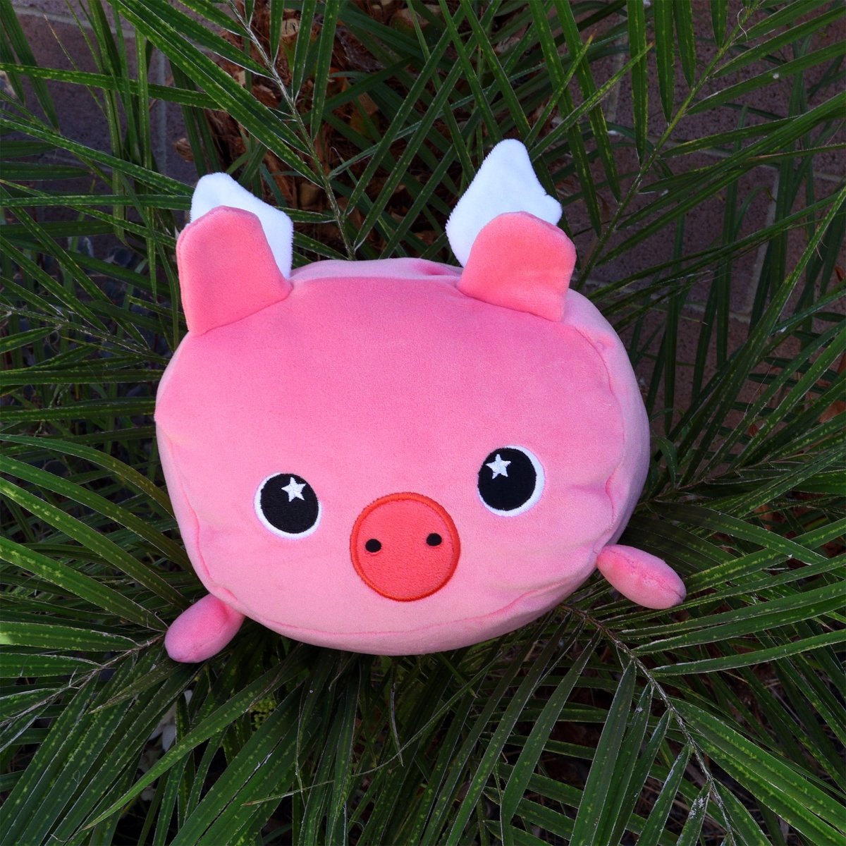 Barnaby the Flying Pig is a round, pink plush toy with white wings and big black eyes, nestled among green palm leaves—a soft, cuddly addition perfect for any collection.