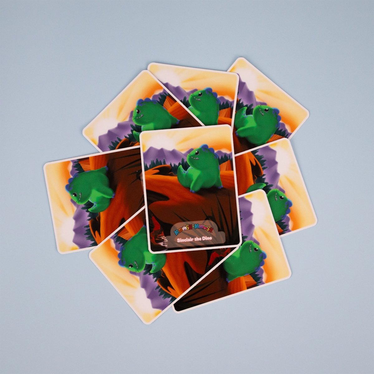 Eight Moosh-Moosh 3" x 4" Vinyl Stickers are arranged in a circle on a light blue surface. Each sticker shows a green dinosaur sitting with mountains and an orange sky behind it. The center sticker says “Snacks the Dino.”.