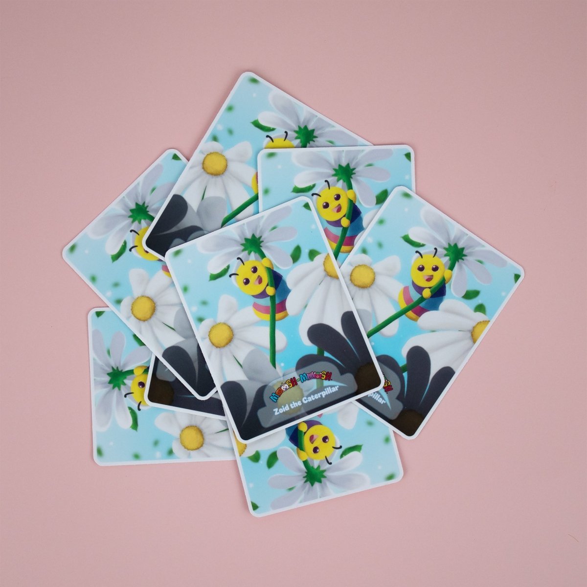Seven colorful Moosh-Moosh 3" x 4" vinyl stickers, each featuring a smiling caterpillar on flowers, are arranged to overlap on a pastel pink background. The bottom sticker reads “Zed the Caterpillar.”.